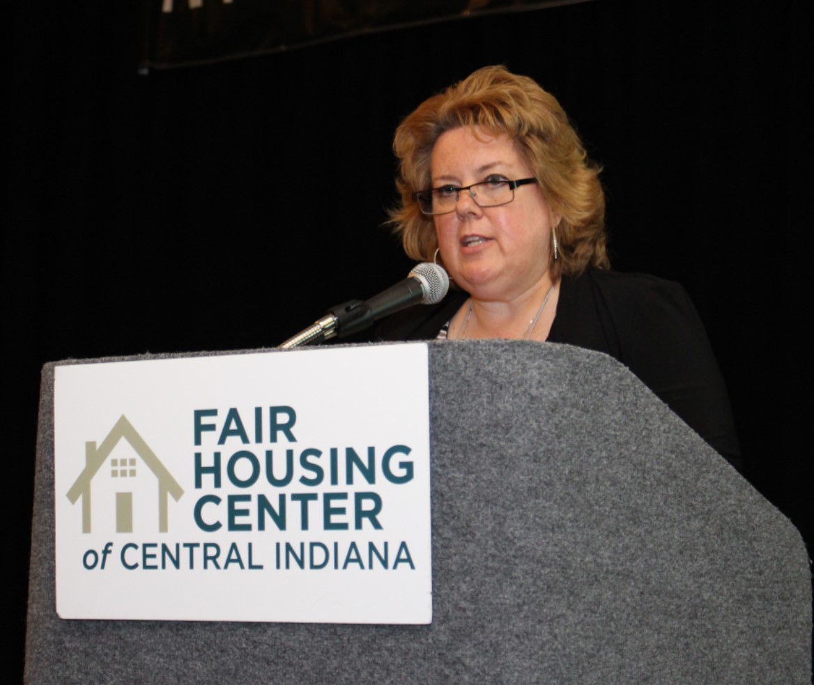 Amy Nelson speaks into a microphone in this photo. She has dark blonde, shoulder-length hair and is wearing glasses.