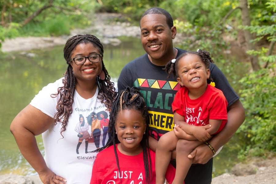 Erica and Aaron Parker smile in this photo with their two daughters. Erica Parker's long hair is in braids, and she's wearing glasses. Aaron Parker is wearing a black t-shirt. He has a mustache and short-cropped hair. Both daughters are smiling and wearing red t-shirts.