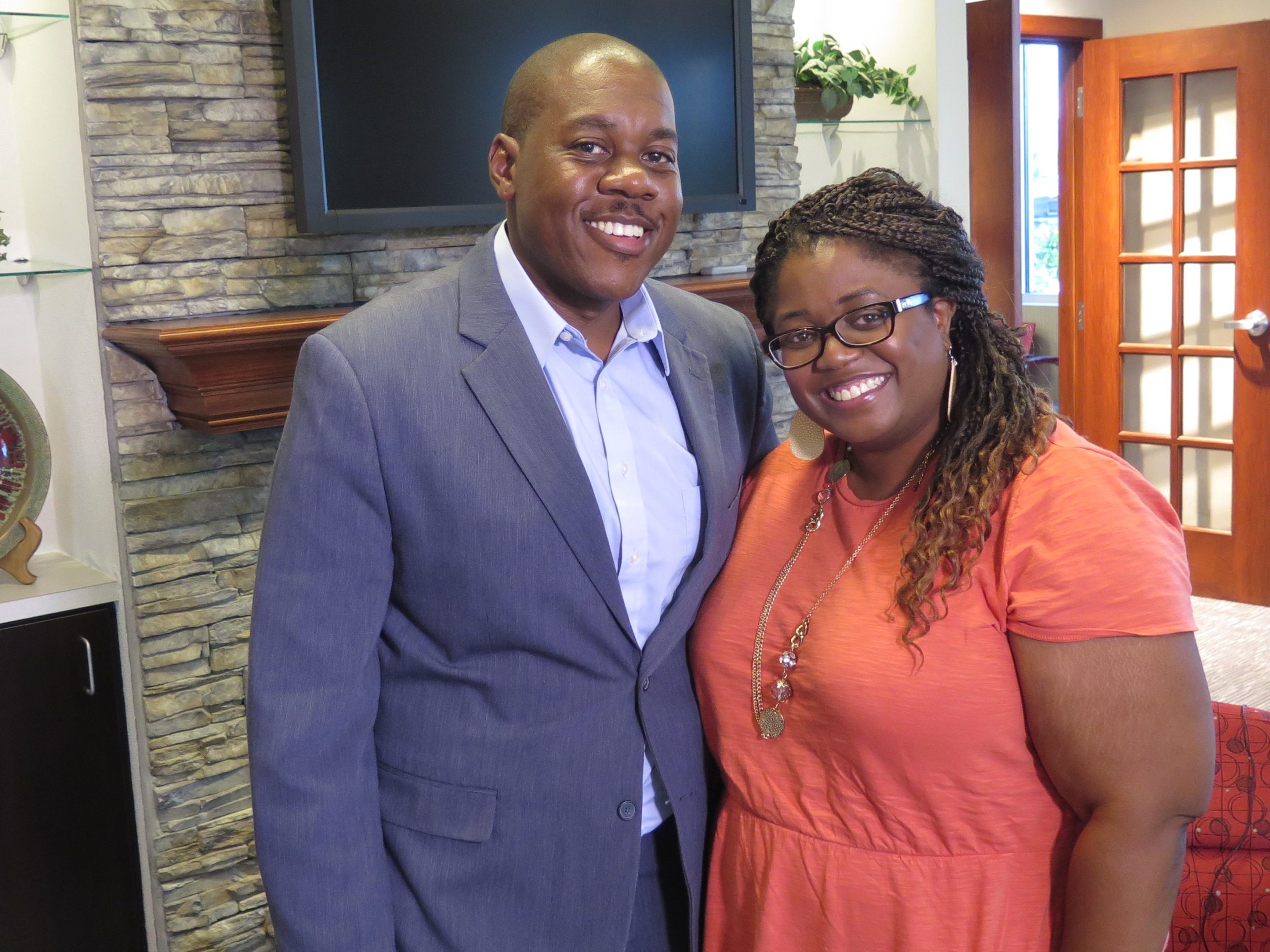 Aaron and Erica Parker pose for a photo at the Sibcy Cline office in West Chester. Aaron Parker is wearing a blue suit and a pale blue dress shirt. His hair is cropped short, and he has a mustache. Erica Parker is wearing a coral colored dress and glasses. Her hair is in long braids.